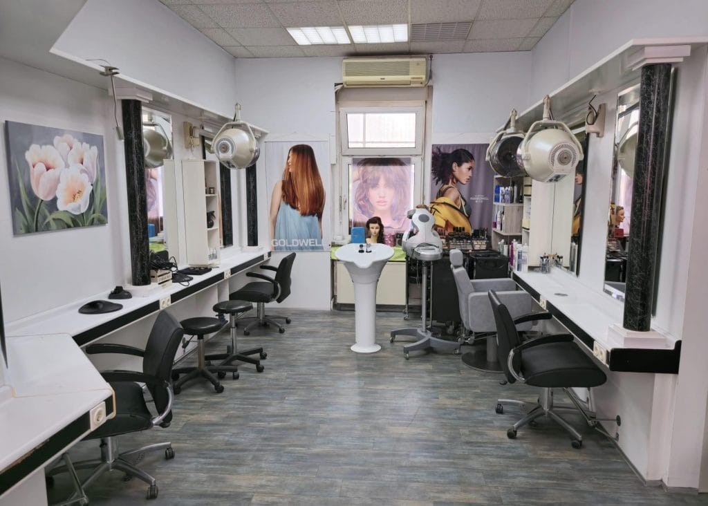 Innenraum eines leeren Friseursalons mit mehreren Frisierplätzen. Jede Station verfügt über einen Stuhl, einen Spiegel und einen Haartrockner. Plakate mit Frisuren schmücken die Wände, und in der Mitte befindet sich eine Auslage mit Haarprodukten. Der Boden ist aus Holz.