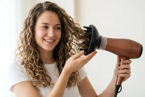 Eine junge Frau mit langen, lockigen braunblonden Haaren trocknet ihre Haare mit einem braunen Föhn, an dem ein schwarzer Remington Universal Diffusor-Aufsatz mit vielen kurzen Stacheln und flexiblem Adapter befestigt ist. Sie trägt ein weißes Shirt, lächelt freundlich mit einem attraktiven, entspannten Gesicht, und die Aufnahme erfolgt aus einer seitlichen Perspektive vor hellem Hintergrund.