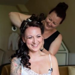 Eine Frau mit gelocktem Haar und einer Tätowierung auf dem Arm lächelt, während eine andere Frau ihr Haar stylt. Beide befinden sich in einem Raum mit warmer Beleuchtung, und die Stylistin sieht konzentriert und glücklich aus.