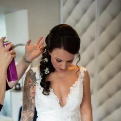 Eine Braut in einem Hochzeitskleid aus Spitze blickt nach unten, während eine Person eine Dose hält und ihr Haar besprüht. Sie trägt ein Haarteil aus Blumen und Spitze, und ihr linker Arm ist mit einer bunten Tätowierung versehen. Im Hintergrund sind gesteppte Paneele an der Wand zu sehen.