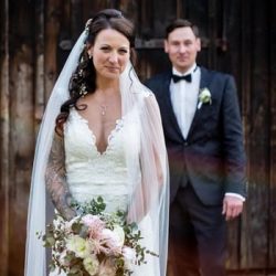 Eine Braut in einem weißen Spitzenkleid hält einen Blumenstrauß und steht vor einem Bräutigam in einem schwarzen Anzug mit Fliege. Sie stehen draußen vor einem rustikalen Holzhintergrund. Die Braut trägt einen Schleier, und beide lächeln.