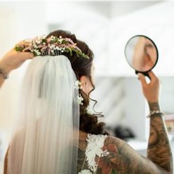Eine Braut mit geblümtem Kopfschmuck und Schleier hält einen kleinen Spiegel, in dem sich ihr Ohr spiegelt. Sie trägt ein Spitzenkleid und hat Tattoos auf dem Arm. Die Umgebung ist hell und weich, möglicherweise eine Umkleidekabine.