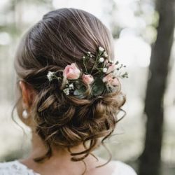 Eine Frau mit braunem Haar, das zu einer eleganten Hochsteckfrisur gestylt ist, geschmückt mit kleinen rosa Rosen und Schleierkraut. Sie steht draußen vor einem verschwommenen grünen Hintergrund und ist in weiße Spitze gekleidet.