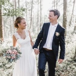 Eine Braut und ein Bräutigam lächeln sich an, während sie sich in einem sonnenbeschienenen Wald an den Händen halten. Die Braut trägt ein weißes Hochzeitskleid aus Spitze und hält einen Strauß aus rosa und weißen Blumen in der Hand. Der Bräutigam trägt einen schwarzen Anzug mit einer rosa Ansteckblume.
